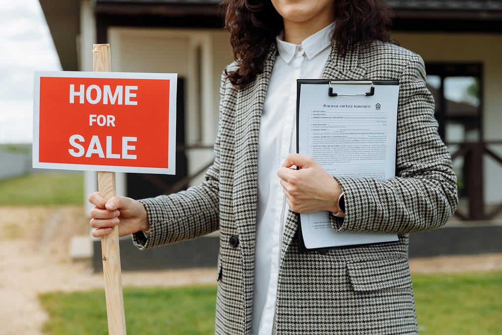 A woman smiles while holding a "For Sale" sign in front of a house, showcasing the Melbourne housing market.